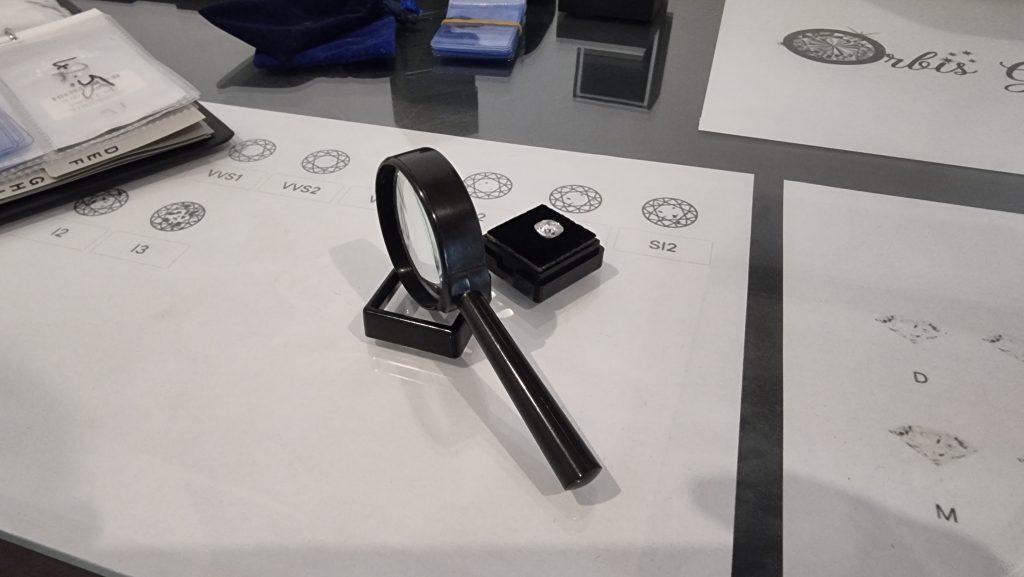 A magnifying loupe rests on a jeweler's grading chart displaying various clarity grades, with a moissanite stone in a box, indicating the precision grading of moissanite gemstones.