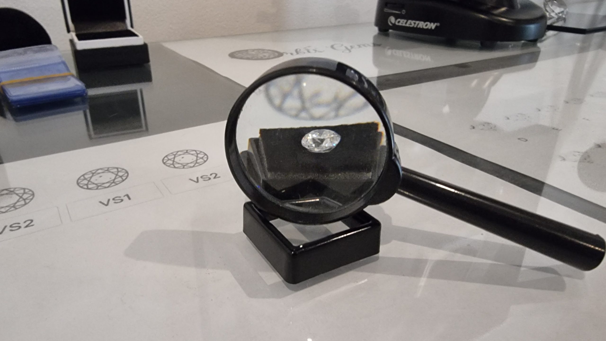 A moissanite stone magnified under a loupe on a jeweler's grading chart, with various clarity grades labeled VS2, VS1.